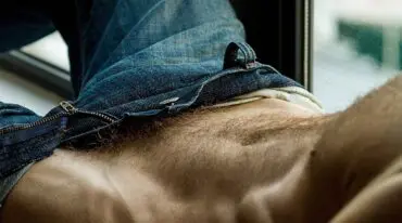 Torso de un hombre adulto recostado con los jeans abiertos y el pubis visible.