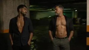 Un hombre joven adulto y un hombre maduro se miran con deseo en un estacionamiento nocturno.