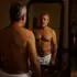 Hombre maduro con el torso desnudo y una toalla frente al espejo de un dormitorio, mientras otro hombre adulto lo observa desde la puerta.
