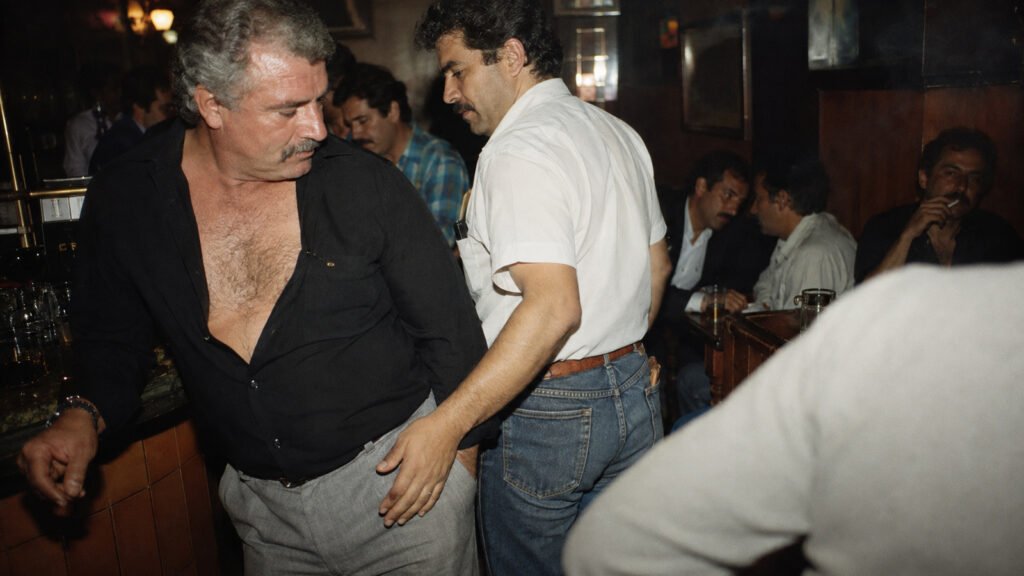 Recreación de hombres adultos que se rozan y observan en un bar gay de finales de los años ochenta o principios de los noventa.