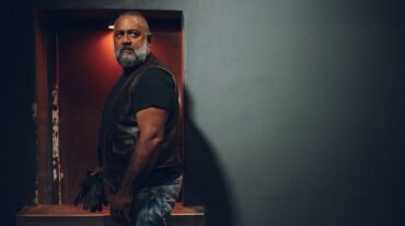 Hombre adulto de barba canosa y chaleco de cuero negro permanece junto a la entrada de un local nocturno.