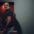 Hombre adulto de barba canosa y chaleco de cuero negro permanece junto a la entrada de un local nocturno.