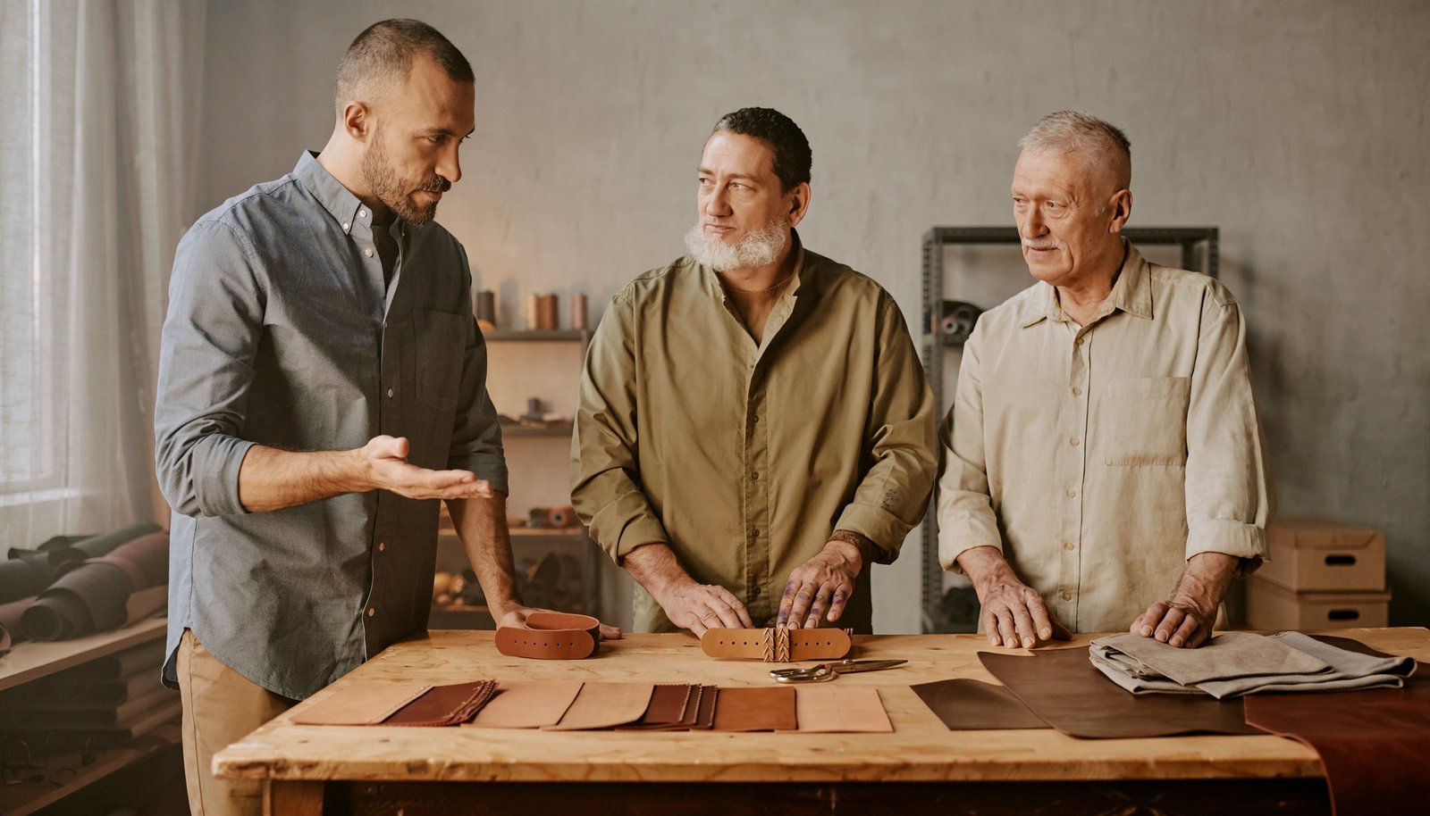 Tres hombres adultos conversan sobre piezas de cuero y materiales dispuestos en una mesa de taller.