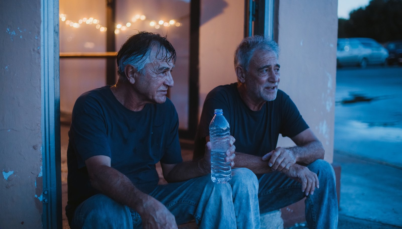 Dos hombres adultos descansan con una botella de agua junto a la salida de un local nocturno al amanecer.