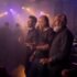 Hombres adultos de distintas edades bailan en un recinto nocturno iluminado con luces violetas.