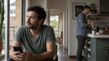 Un hombre adulto mira hacia la ventana mientras su pareja permanece en la cocina; escena editorial sobre confianza en pareja.