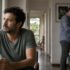 Un hombre adulto mira hacia la ventana mientras su pareja permanece en la cocina; escena editorial sobre confianza en pareja.