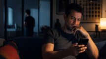 Hombre adulto sentado en un sofá oscuro mirando su teléfono; otra figura aparece desenfocada detrás.