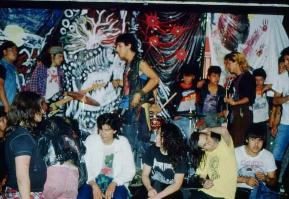 Banda musical con bajo, guitarra y batería frente a un mural colorido; varias personas escuchan y conversan junto al escenario.