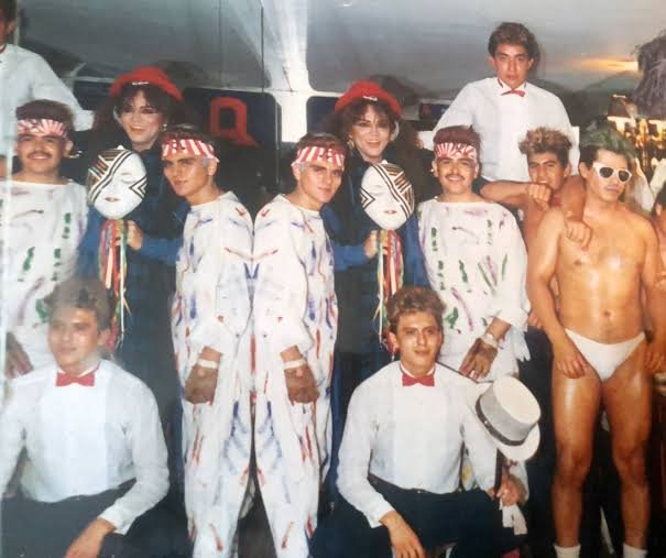 Grupo de personas posa con vestuario de espectáculo, camisas blancas y ropa formal en un interior con espejos.