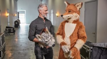 Un hombre adulto sostiene una cabeza de disfraz de lobo junto a otro adulto vestido con un traje de zorro en un corredor.