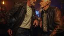 Dos hombres maduros con canas conversan sonrientes en un bar cálido; uno sostiene un pañuelo de tela azul junto a su chamarra de cuero.