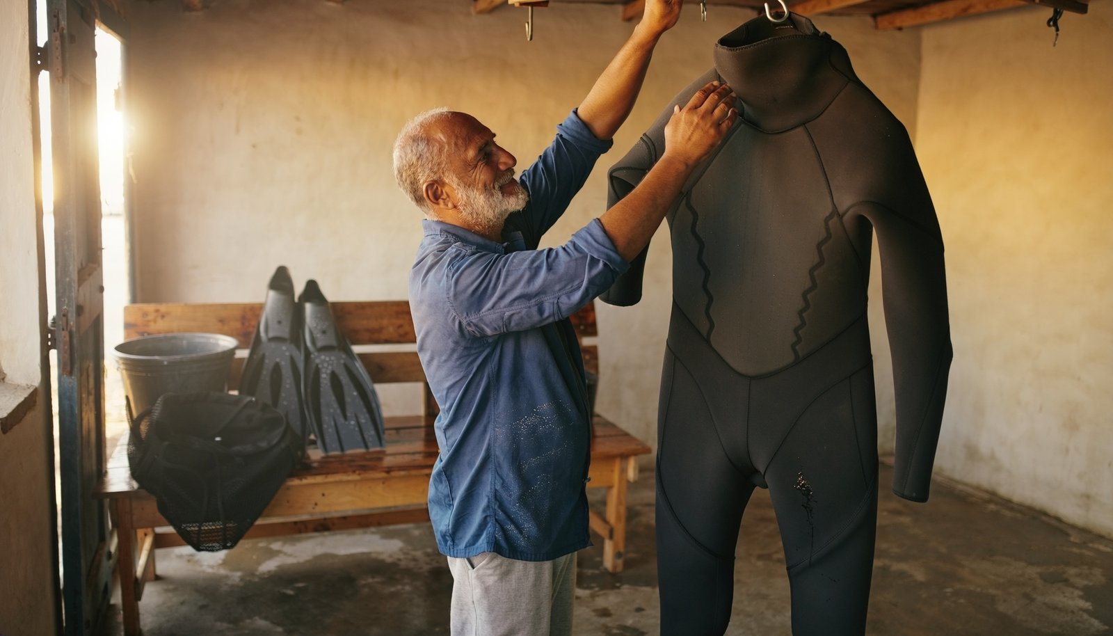 Hombre adulto de barba canosa cuelga un traje de neopreno negro para secarlo en un taller de equipo acuático.