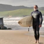 Surfista mexicano adulto de barba canosa, vestido con traje de neopreno, sostiene una tabla junto a las olas de una playa del Pacífico.