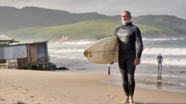 Surfista mexicano adulto de barba canosa, vestido con traje de neopreno, sostiene una tabla junto a las olas de una playa del Pacífico.