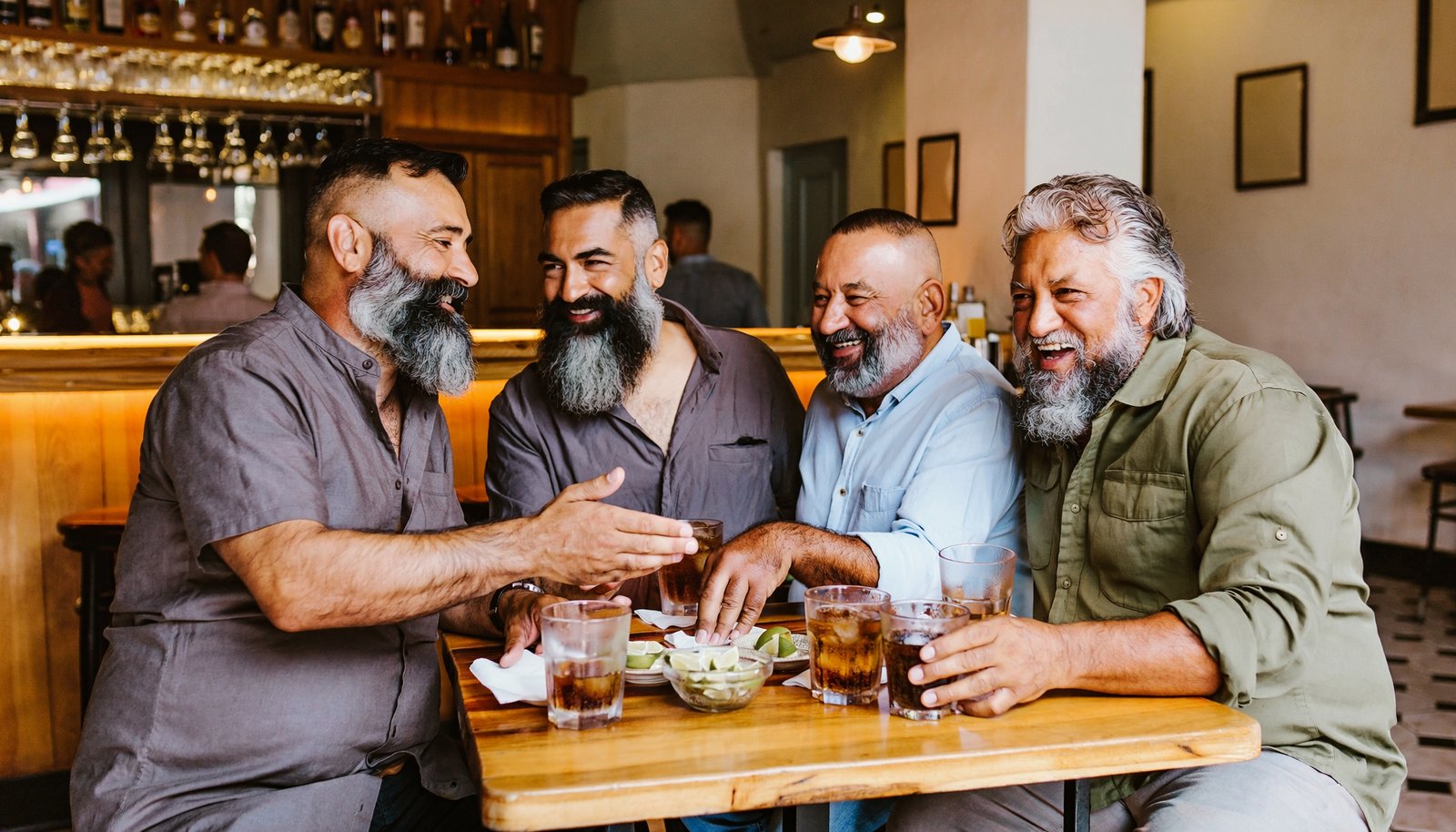 Cuatro hombres mexicanos adultos, de barba y distintas complexiones, conversan y ríen alrededor de una mesa en un bar.