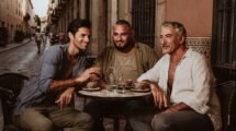 Tres hombres adultos de distintas edades y complexiones conversan en una mesa de café al aire libre.