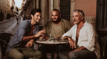 Tres hombres adultos de distintas edades y complexiones conversan en una mesa de café al aire libre.