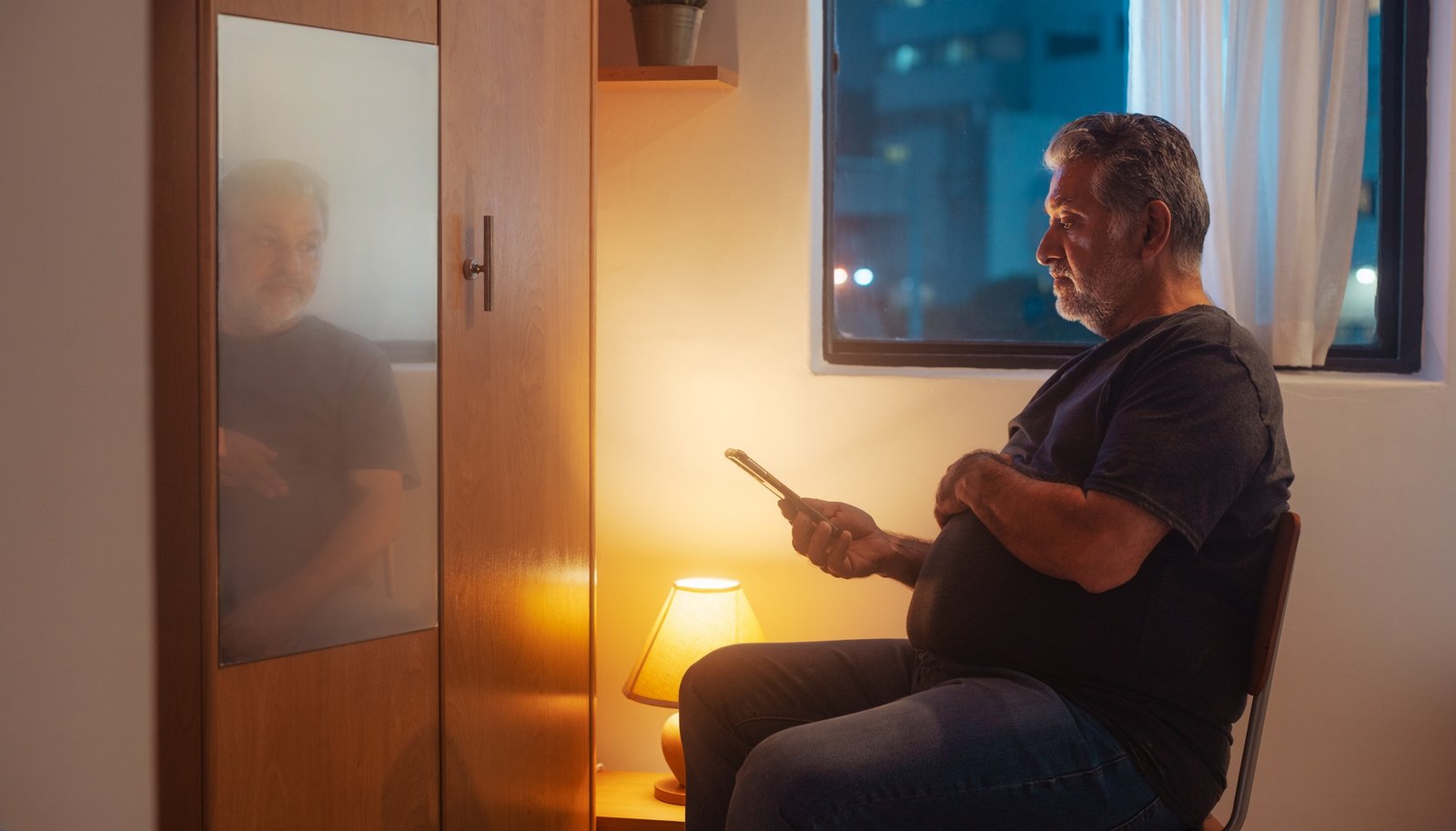 Hombre adulto maduro observa su teléfono mientras su reflejo aparece en el espejo de una habitación.