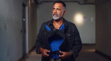 Hombre adulto de barba canosa sostiene una máscara de juego canino azul y negra en un pasillo.