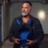 Hombre adulto de barba canosa sostiene una máscara de juego canino azul y negra en un pasillo.