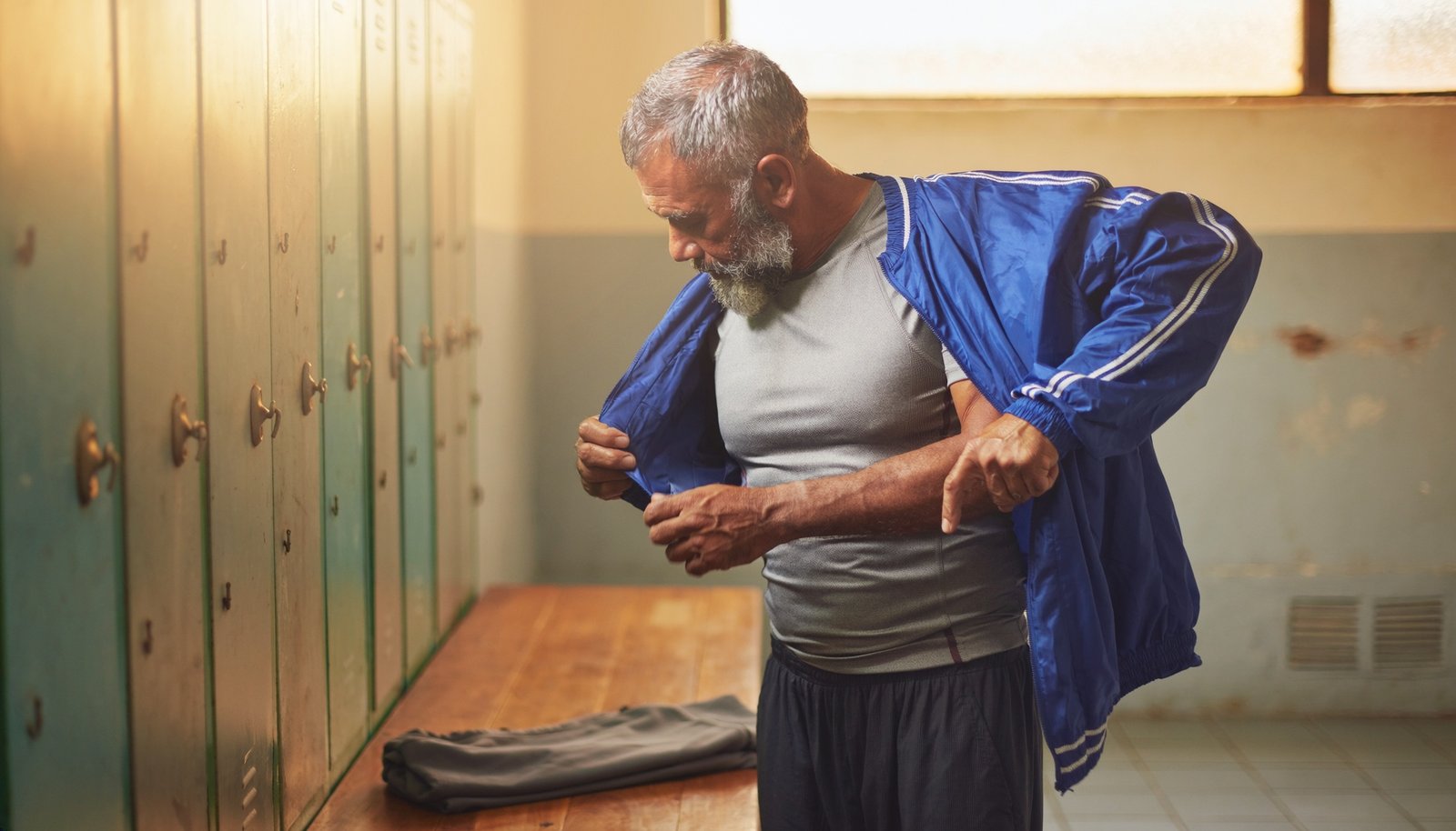 Hombre mexicano adulto de barba canosa se ajusta una chamarra deportiva azul junto a los casilleros de un vestuario.