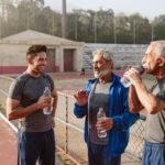 Tres hombres mexicanos adultos con ropa deportiva conversan y beben agua junto a la pista de entrenamiento de un parque.