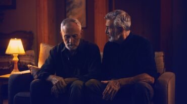 Dos hombres adultos de cabello y barba canosos permanecen sentados juntos en un sofá, uno reflexivo y el otro atento, en una habitación iluminada por una lámpara cálida.
