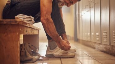 Hombre mexicano adulto de barba canosa se inclina para atar los cordones de unos tenis gastados en un vestuario con casilleros.