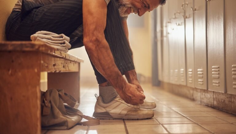 Hombre mexicano adulto de barba canosa se inclina para atar los cordones de unos tenis gastados en un vestuario con casilleros.